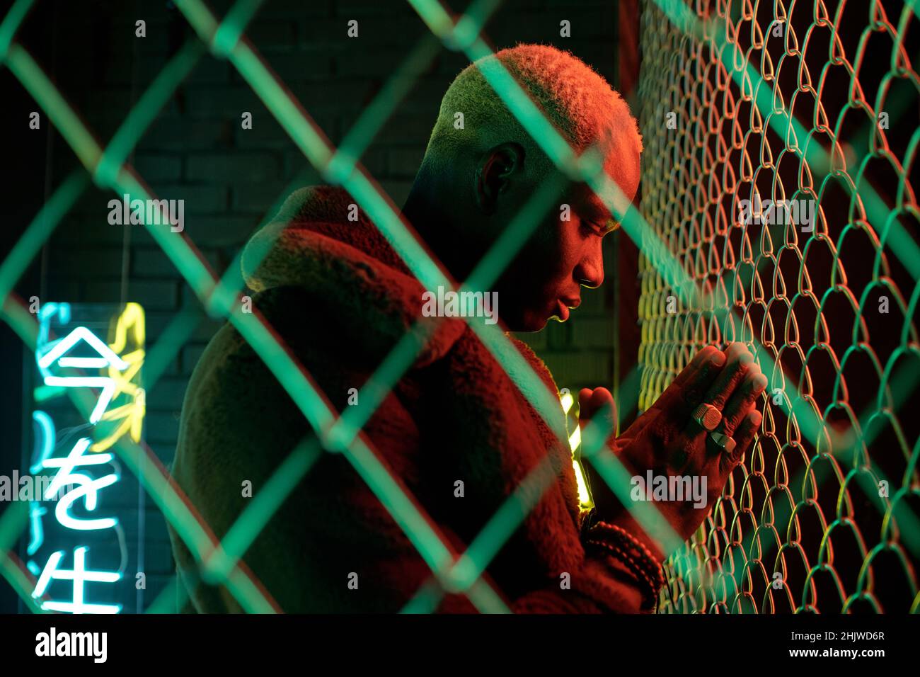 Afrikanischer junger Mann im Pelzmantel, der sich mit den Händen vor dem Gesicht verbeugt, während er an Gittern in Verstecken steht Stockfoto