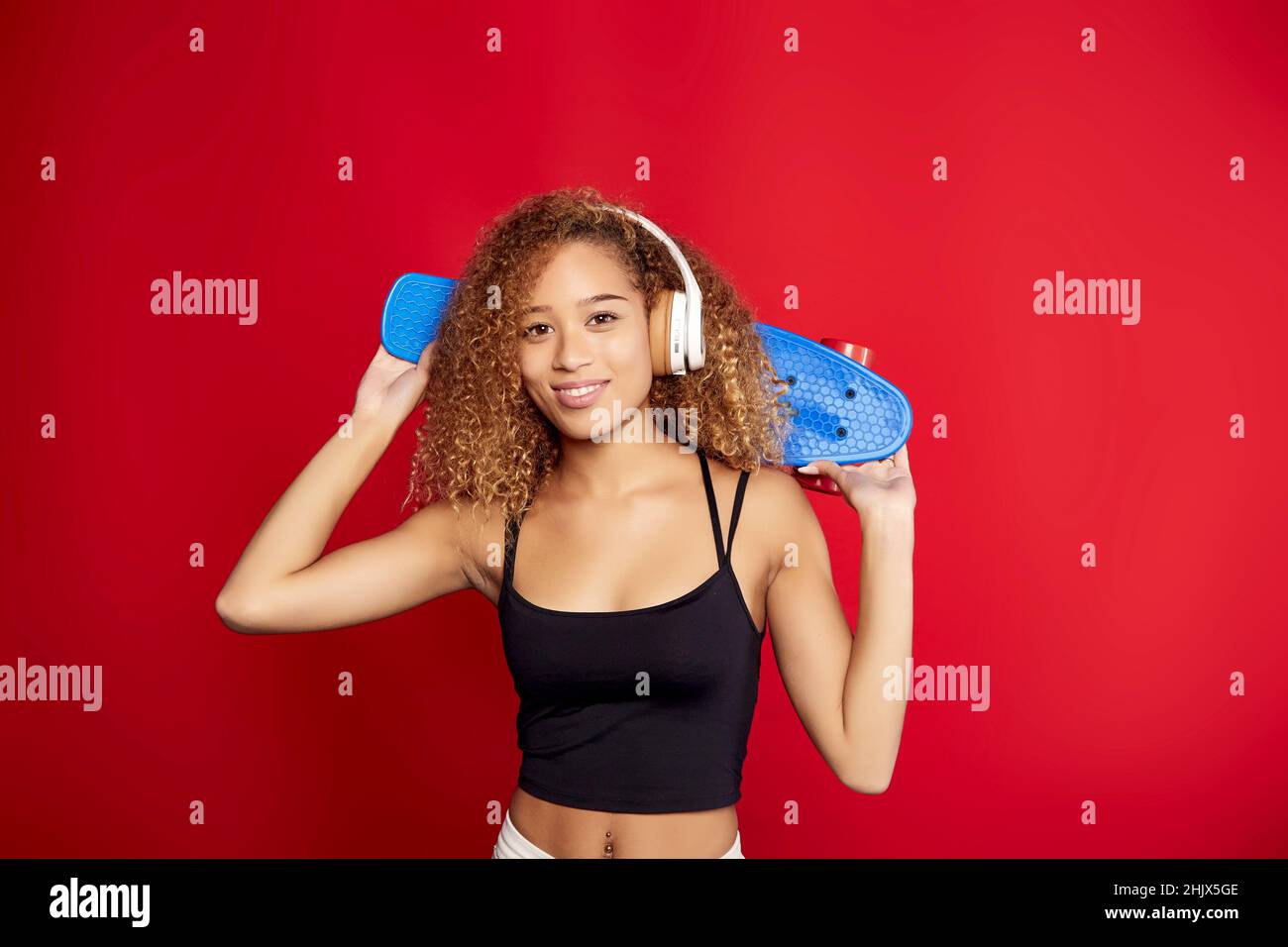 Fröhliche afroamerikanische Hipsterin mit lockigen Haaren mit Penny Board, die Musik in Kopfhörern hört und im Studio vor rotem Hintergrund auf die Kamera schaut Stockfoto