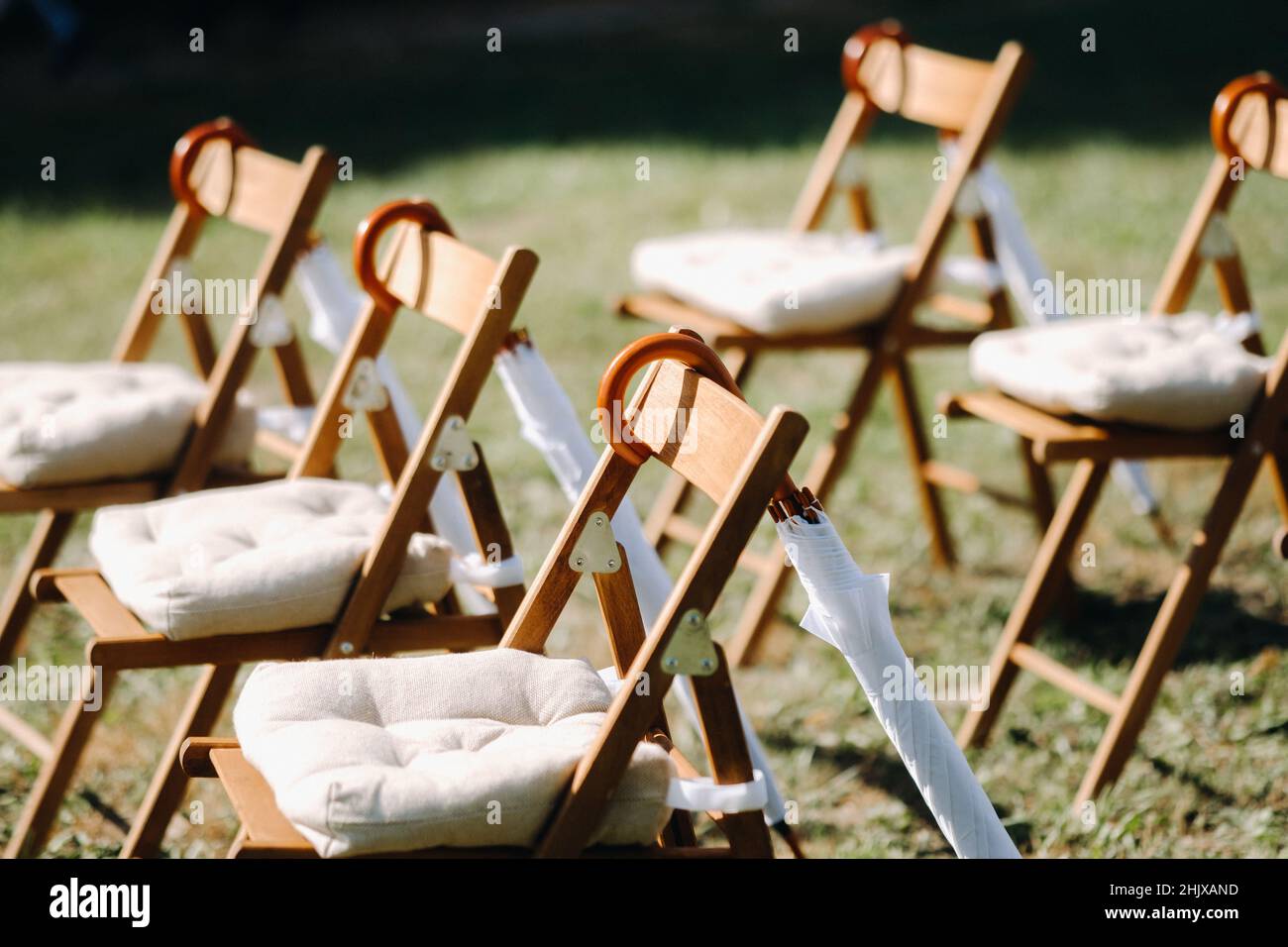 Stühle stehen auf dem grünen Gras im Bereich der Hochzeitszeremonie, weiße Schirme hängen auf den Stühlen. Stockfoto