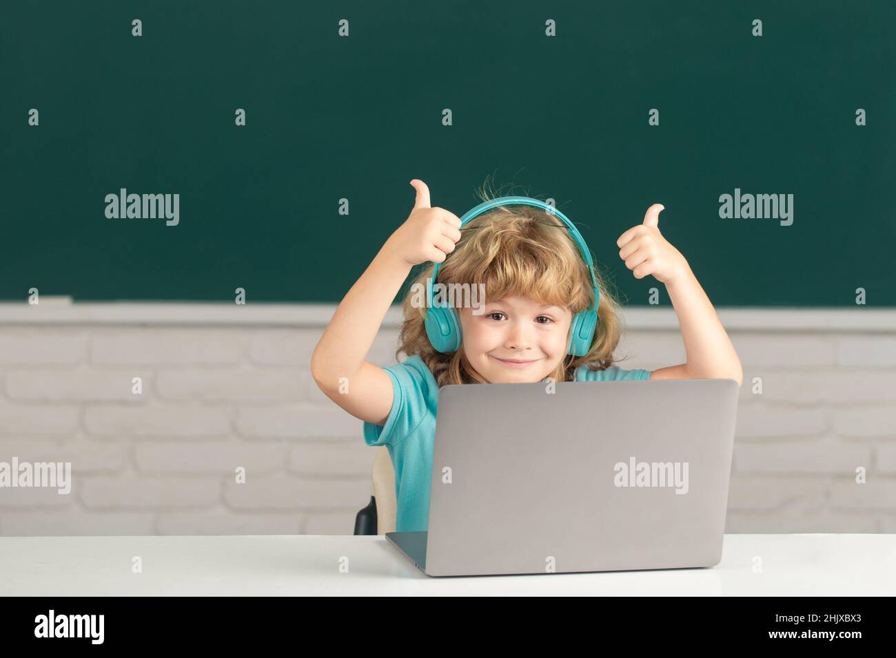 Cleverer Schuljunge mit Daumen nach oben, niedlicher Schüler trägt Kopfhörer und schreibt auf dem Laptop, hört Audiounterricht am Computer. Videocall, E-Study mit Tutor Stockfoto