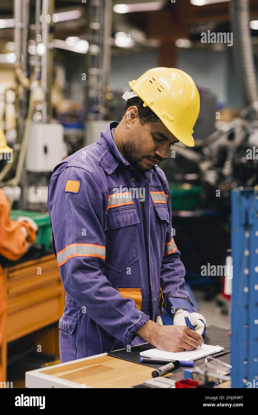 Männlicher Arbeiter mit blauem Kragen, der in Notizblock schreibt, während er in der Industrie arbeitet Stockfoto
