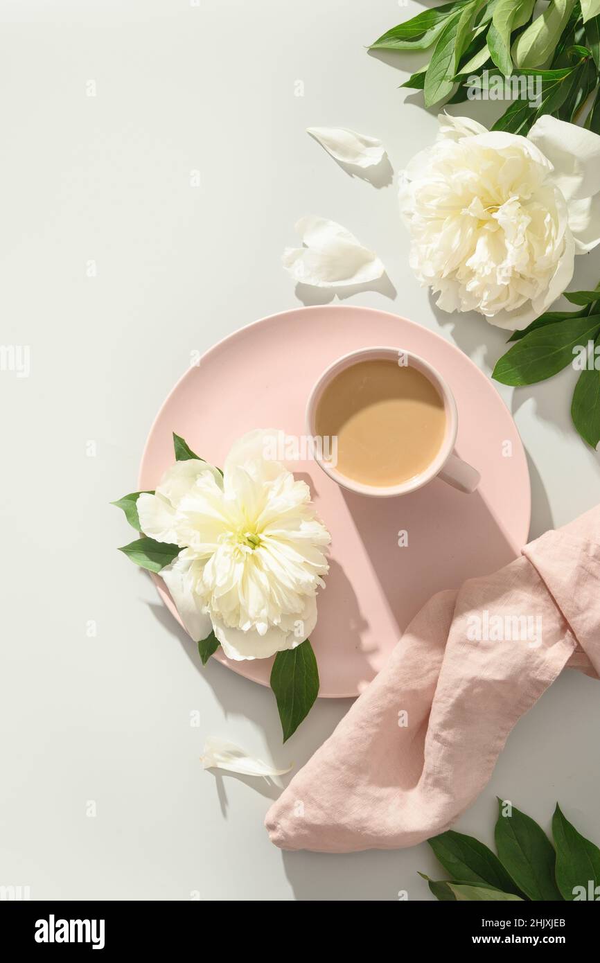 Sommer- oder Frühlingsfrühstück mit Kaffee, weißen Pfingstrosen-Blumen, Bastelhülle auf grauem Hintergrund mit sonnigem Schatten. Vertikaler Arbeitsplatz im Frühling, Top vie Stockfoto