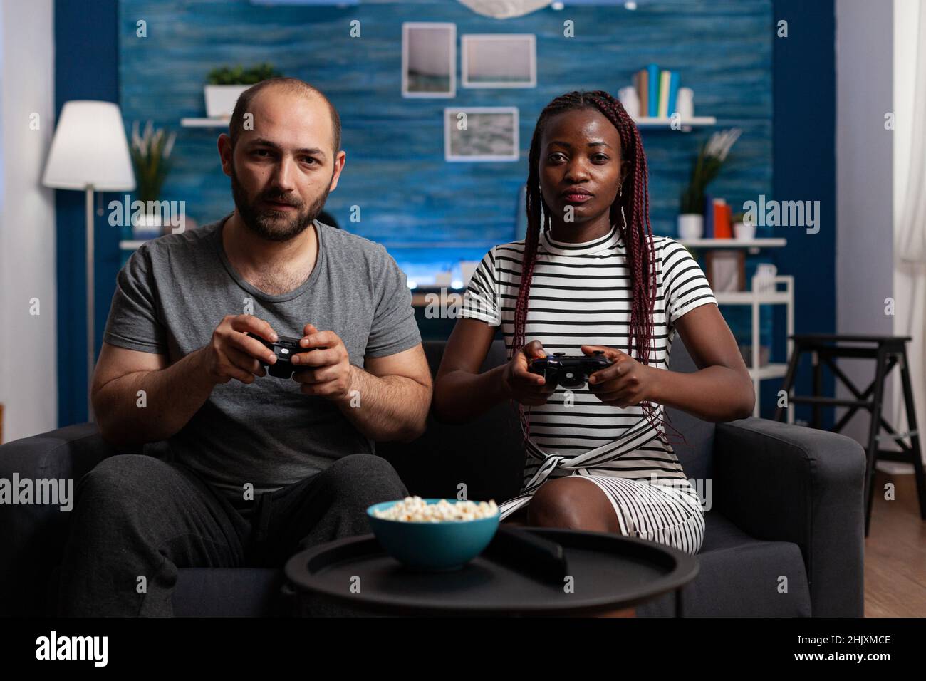 Verheiratetes interrassisches Paar, das Videospiele mit Controllern spielt, während es zu Hause auf dem Sofa sitzt. Entspannte Erwachsene genießen Konsolenspiele und Unterhaltungsaktivitäten zusammen Stockfoto