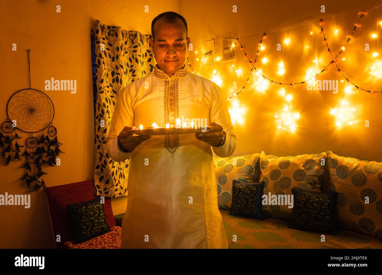 Junger Mann in traditioneller Kleidung, der die Plate mit Öllampen mit Fährenbeleuchtung im Hintergrund hält Stockfoto
