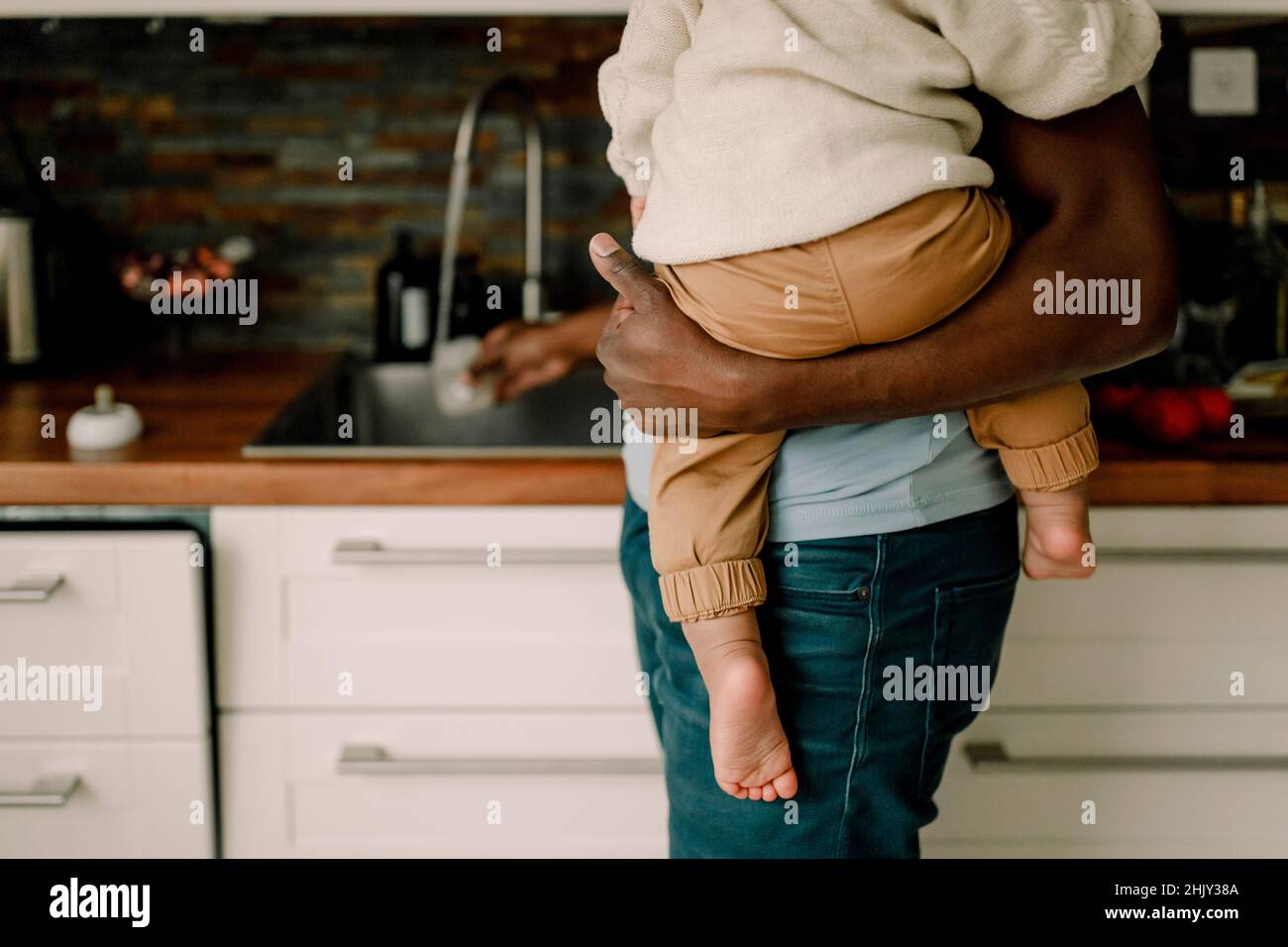 Mittelteil des Vaters, der einen kleinen Jungen trägt, während er in der Küche Aufgaben erledigt Stockfoto