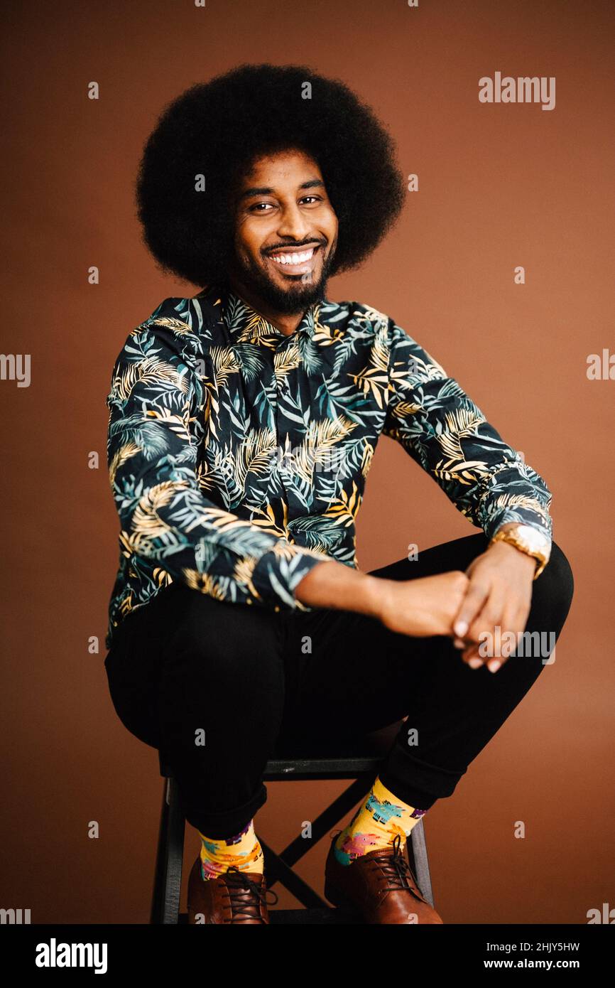 Porträt eines lächelnden Mannes, der auf einem Stuhl vor braunem Hintergrund sitzt Stockfoto