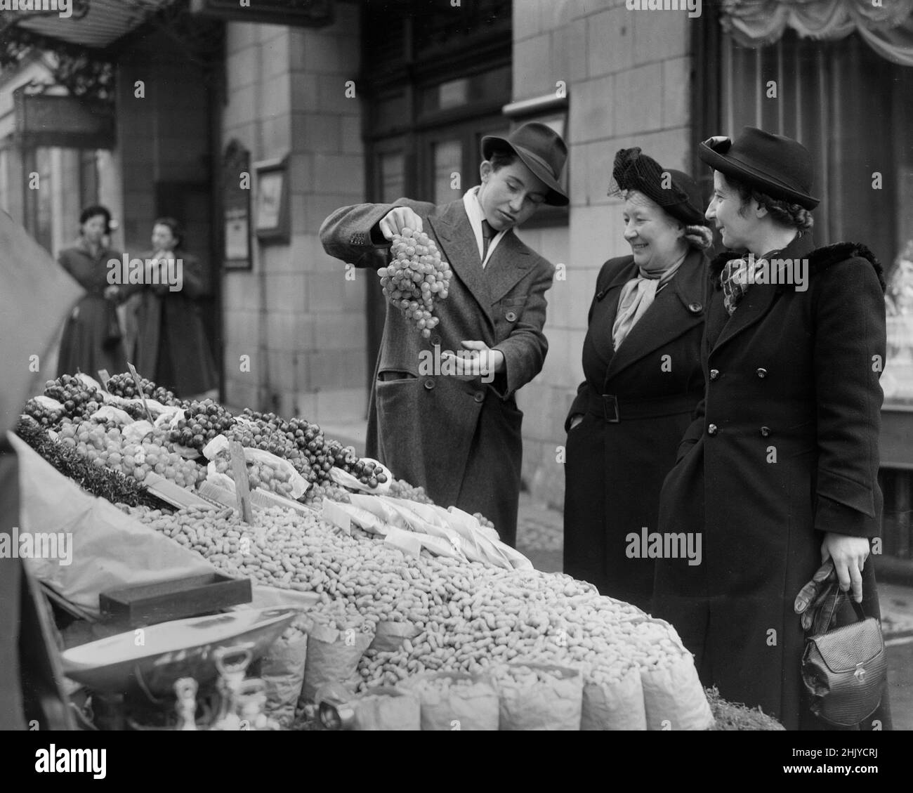 LONDON: Ein Barrow-Junge verkauft in den 1940er Jahren an zwei Frauen mittleren Alters einen Traubenstrauch von seinem Obst- und Gemüsestände am Londoner Strand. Quelle: The DL Archive Collection/Alamy Stockfoto