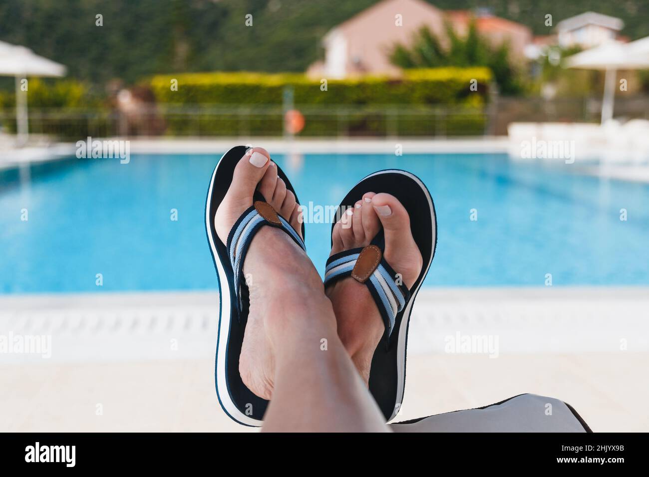 Nahaufnahme der Füße einer jungen Frau in blauen Flip-Flops, die auf einer Sonnenliege mit Swimmingpool im Hintergrund liegen. Kefalonia Island, Griechenland Stockfoto