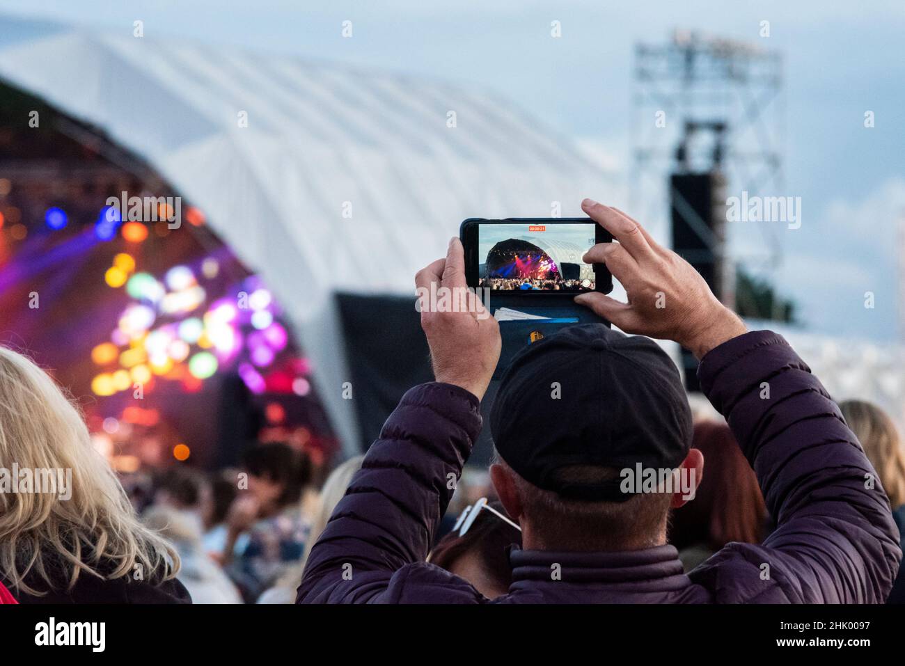 Eine Person, die ein Mobiltelefon benutzt, um die Band zu videospielen, die auf der Bühne eines großen Musikfestivals in einem Park in Essex spielt. Aufzeichnung der Show Stockfoto