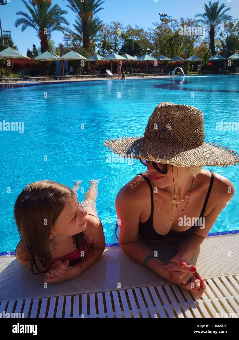 Glückliches hübsches kleines Mädchen mit Mutter im großen Strohhut, als sie sich am Pool des Spa Hotels ausruhen Stockfoto