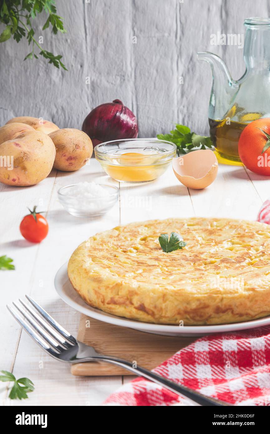 Traditionelles spanisches Omelett mit Kartoffeln und Zwiebeln mit Zutaten auf einem hellen, rustikalen Hintergrund, vertikales Format. Spanische Tapas. Stockfoto