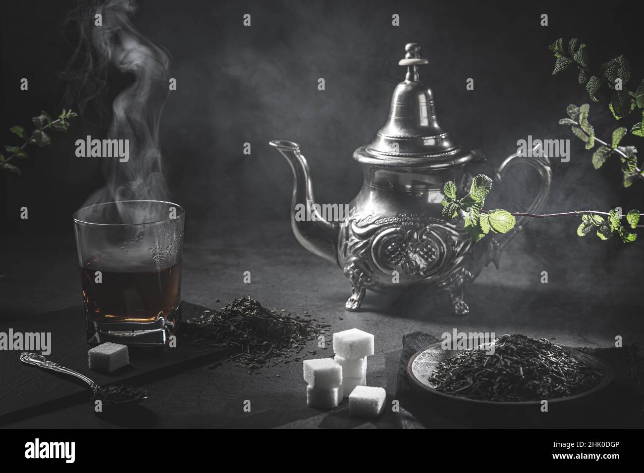 Traditionelle marokkanische Teekane mit einer dampfenden Tasse Tee, Zucker und Minze in rauchiger schwarzer Atmosphäre. Marokkanisches Teekonzept. Stockfoto