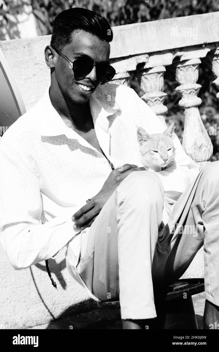 Graustufen eines südasiatischen Mannes in einer Brille, der mit einer Katze im Freien sitzt Stockfoto