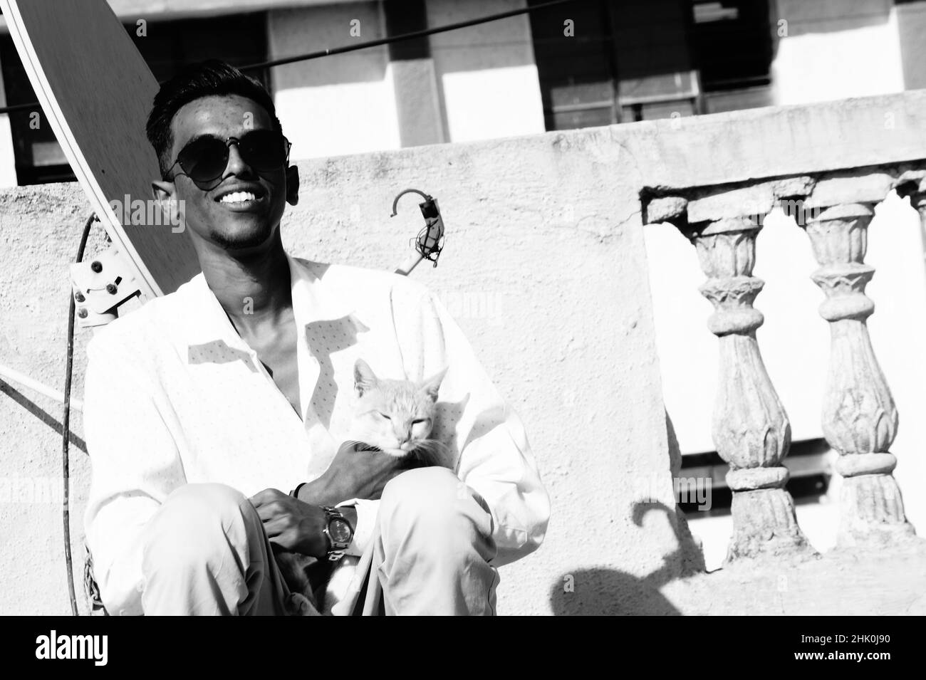 Graustufen eines südasiatischen Mannes in einer Brille, der mit einer Katze im Freien sitzt Stockfoto