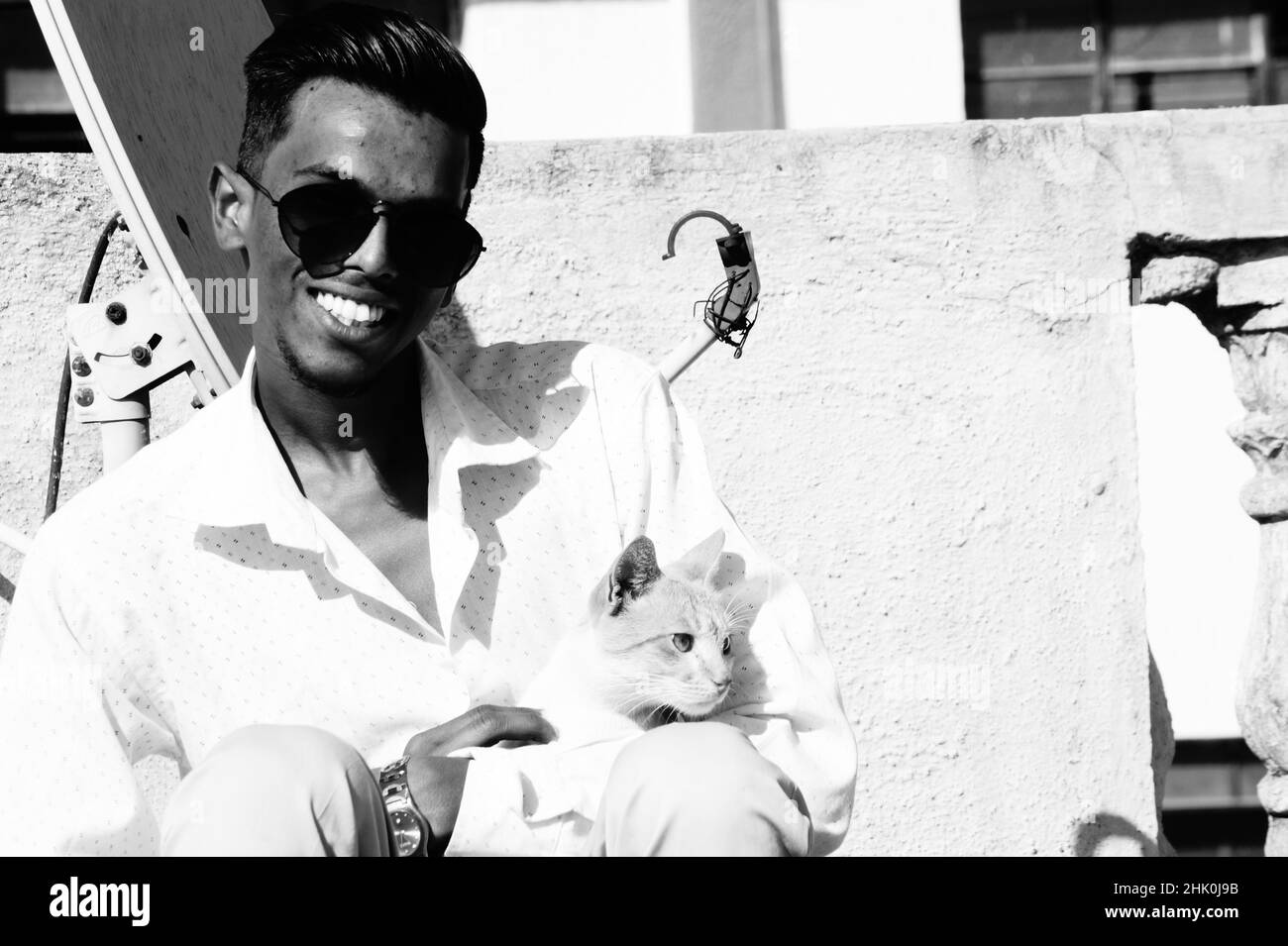 Graustufen eines südasiatischen Mannes in einer Brille, der mit einer Katze im Freien sitzt Stockfoto