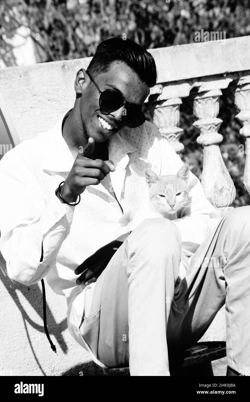 Graustufen eines südasiatischen Mannes in einer Brille, der mit einer Katze im Freien sitzt Stockfoto