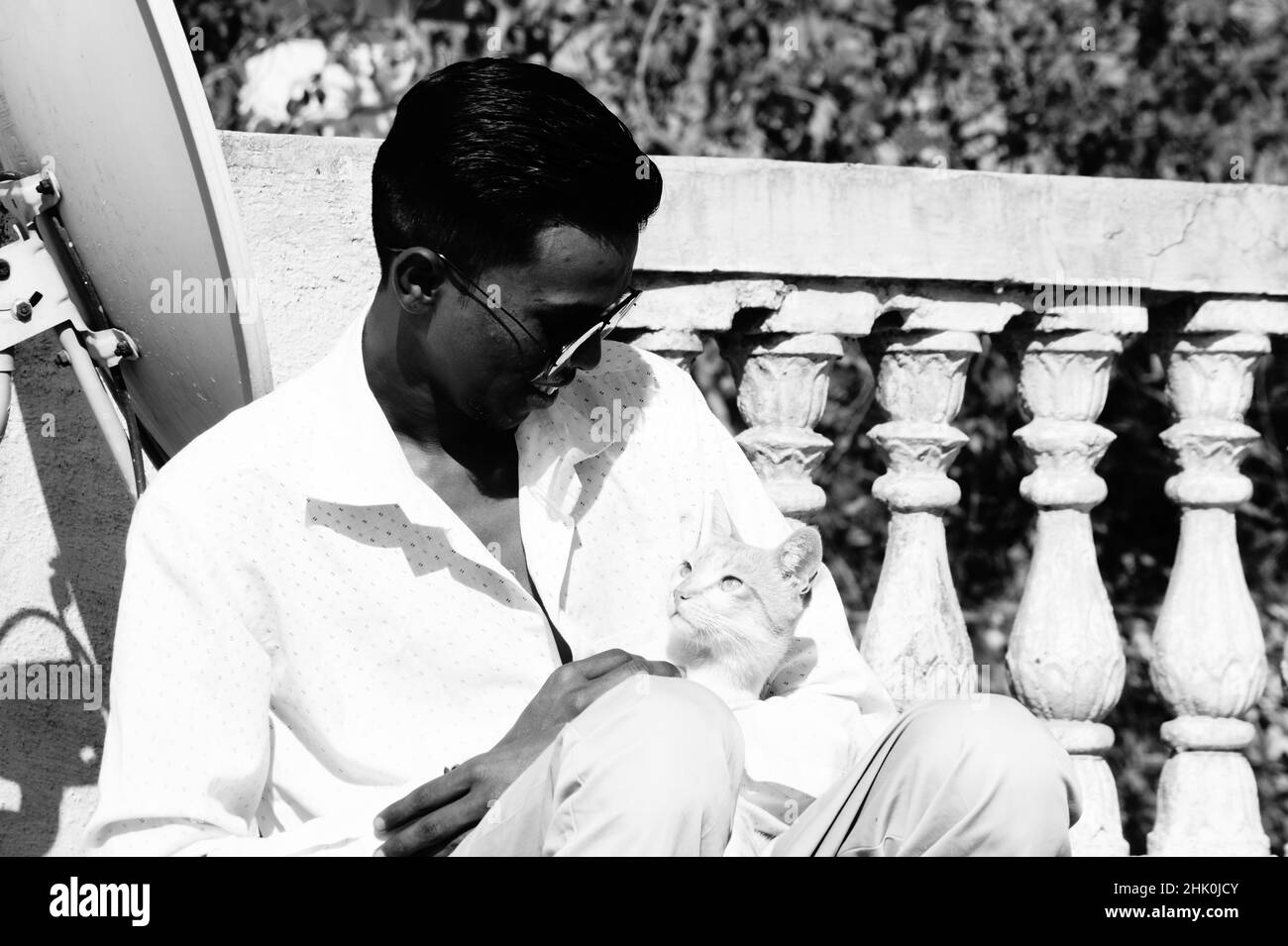 Graustufen eines südasiatischen Mannes in einer Brille, der mit einer Katze im Freien sitzt Stockfoto