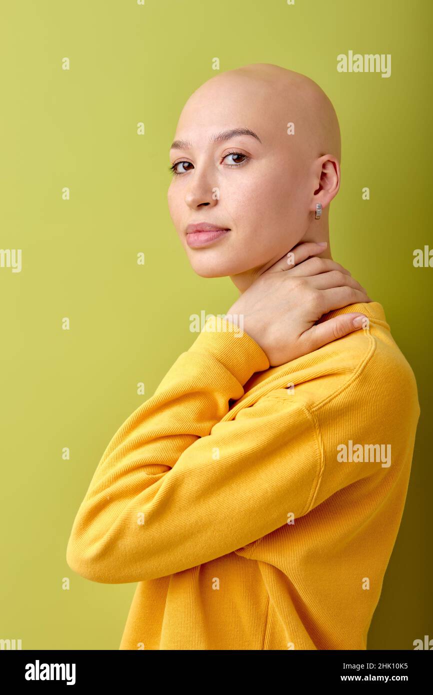 Attraktive Glatze kaukasische Frau in gelbem Hemd stehend isoliert über grünem Studio-Hintergrund, posiert vor der Kamera, berühren Hals. Schöne Dame mit einem Stockfoto