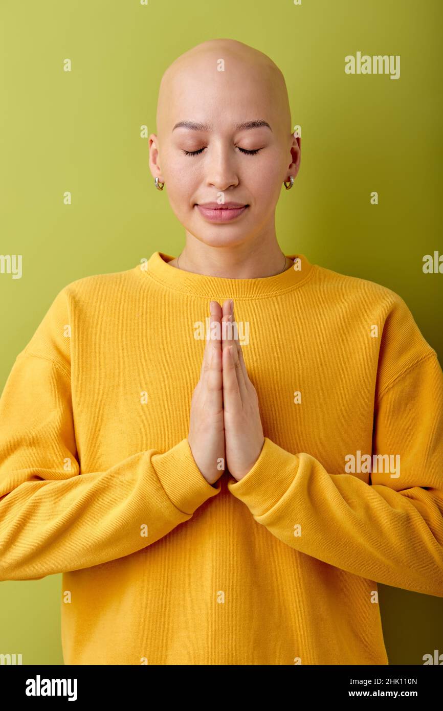 Ruhige und wunderschöne junge Glatze kaukasische Frau mit geschlossenen Augen posiert vor der Kamera und betet. Porträt einer hübschen Dame in gelbem Casual Shirt isoliert über Stockfoto