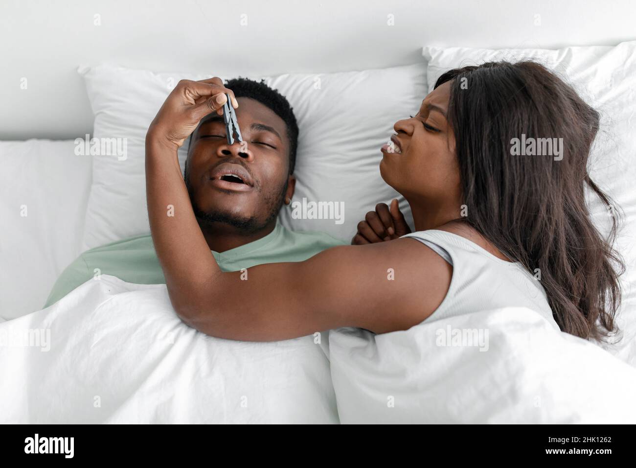 Unzufriedene lustige Millennial schwarze Dame aufwachen aus schnarchen schlafenden Mann, schließt seine Nase mit Wäscheklammer Stockfoto