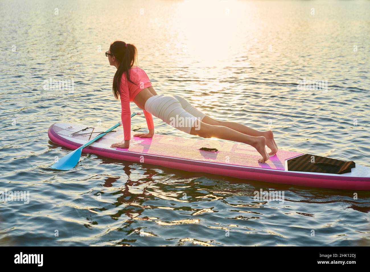 Junge athletische Frau in Sportkleidung, die Plank-Übung auf dem Paddle-Board macht. Schöner Sommeruntergang im Hintergrund. Konzept des Outdoor-Workouts. Stockfoto