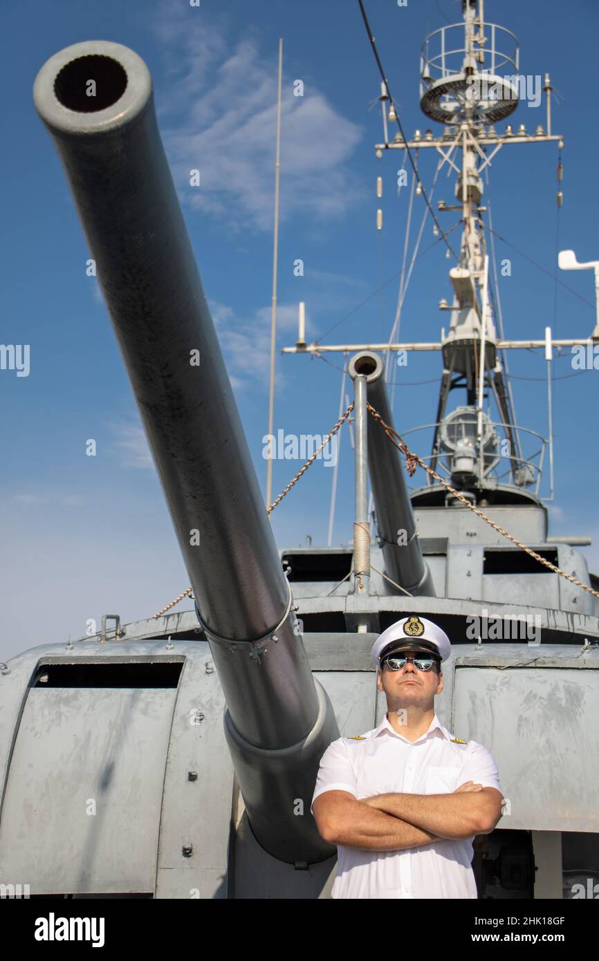 Ein Marineoffizier, der unter der Kanone eines Schiffes steht.der Kapitän in weißer Uniform steht an Bord eines Schiffes. Stockfoto