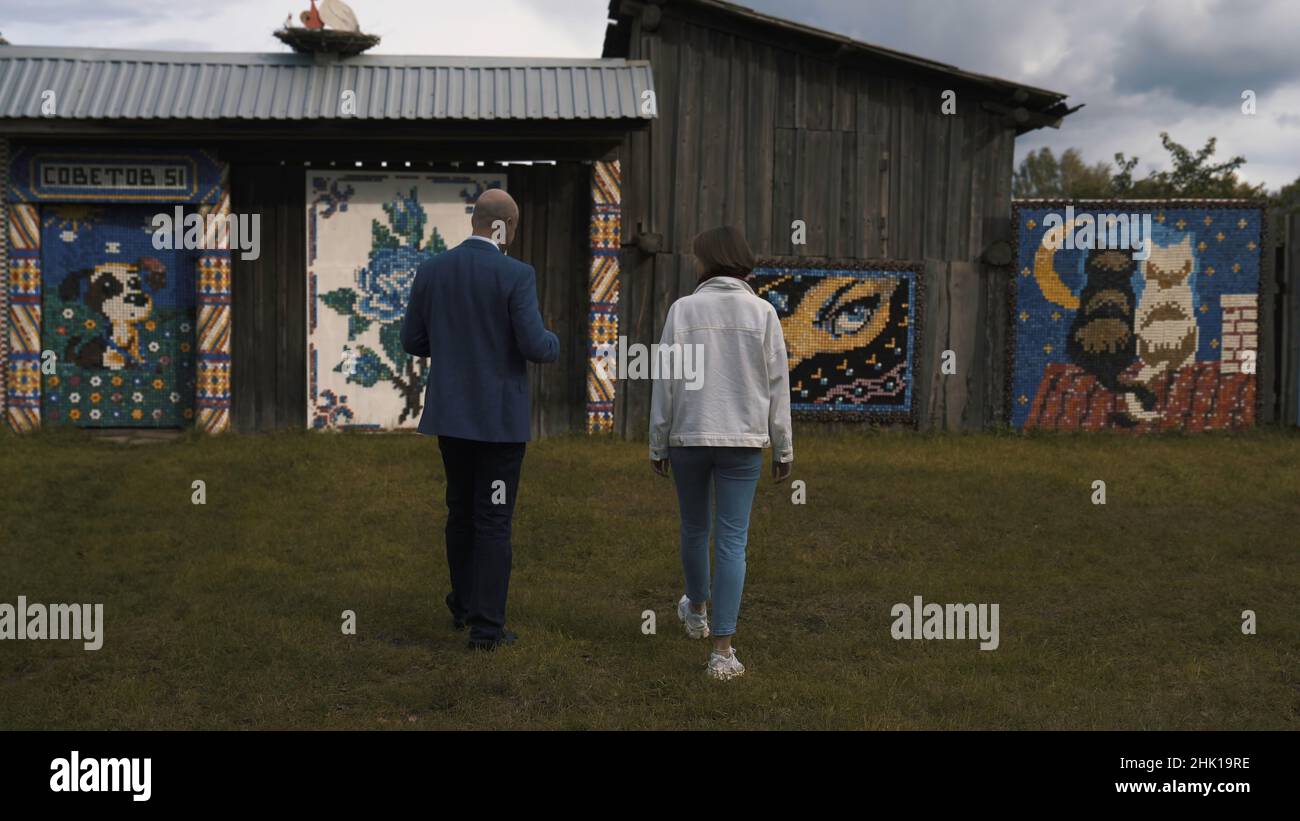 Rückansicht eines Mannes in dunkelblauem Anzug und einer Frau in weißer Jacke und blauer Jeans, der an Holzhäusern mit Mosaikwänden entlang geht. Wände mit wunderschönen MOS Stockfoto