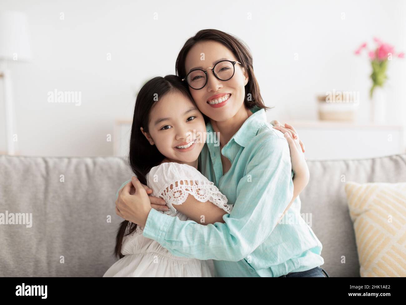 Glückliches kleines asiatisches Mädchen und ihre liebevolle Großmutter umarmen und betrachten die Kamera auf der Couch zu Hause Stockfoto