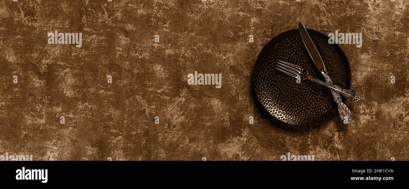 Messer und Gabel quer auf einer Platte. Elegantes Vintage-Messer und Gabel auf einem schwarzen Essteller über texturiertem Hintergrund gekreuzt. Sepia-getöntes breites Bild. Stockfoto