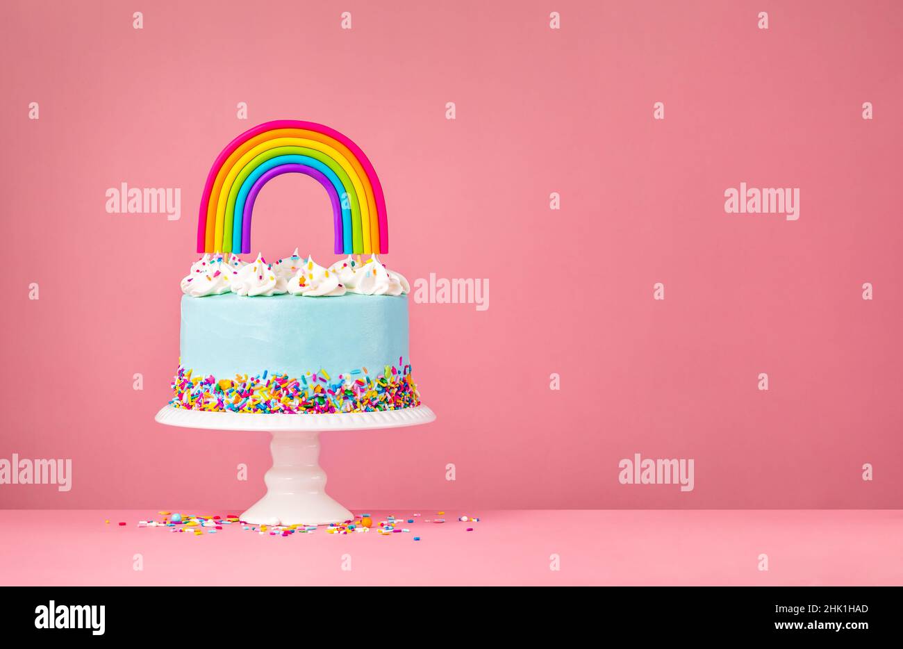 Blauer Geburtstagstorte mit Baiser-Dekorationen, Streuseln und einem farbenfrohen, hausgemachten Regenbogentopfer auf einem rosa Hintergrund. Stockfoto