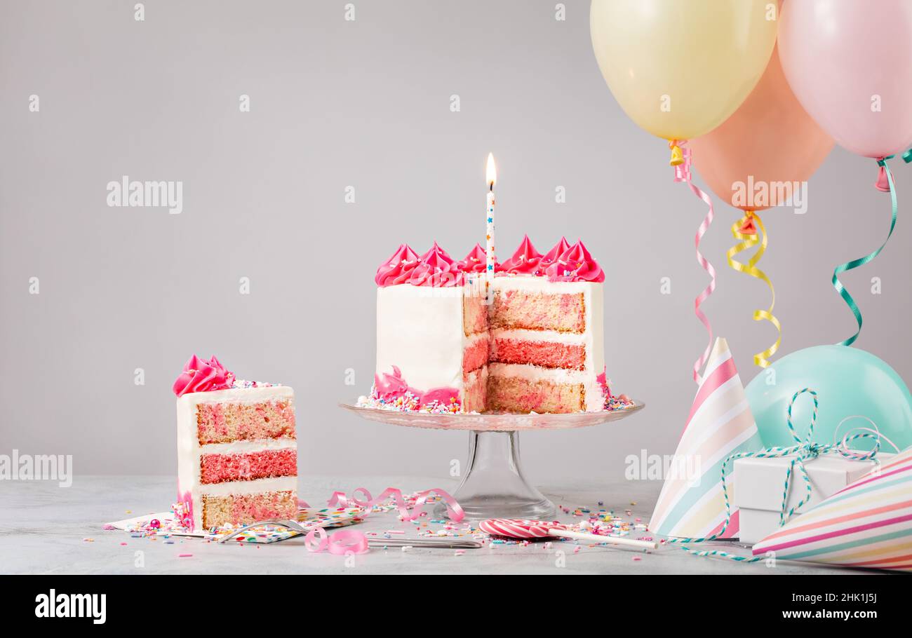 Pinkfarbene Funfetti-Geburtstagstorte mit Kerze, Geschenken, Hüten und bunten Luftballons auf hellgrauem Hintergrund. Szene von einer Geburtstagsparty! Stockfoto