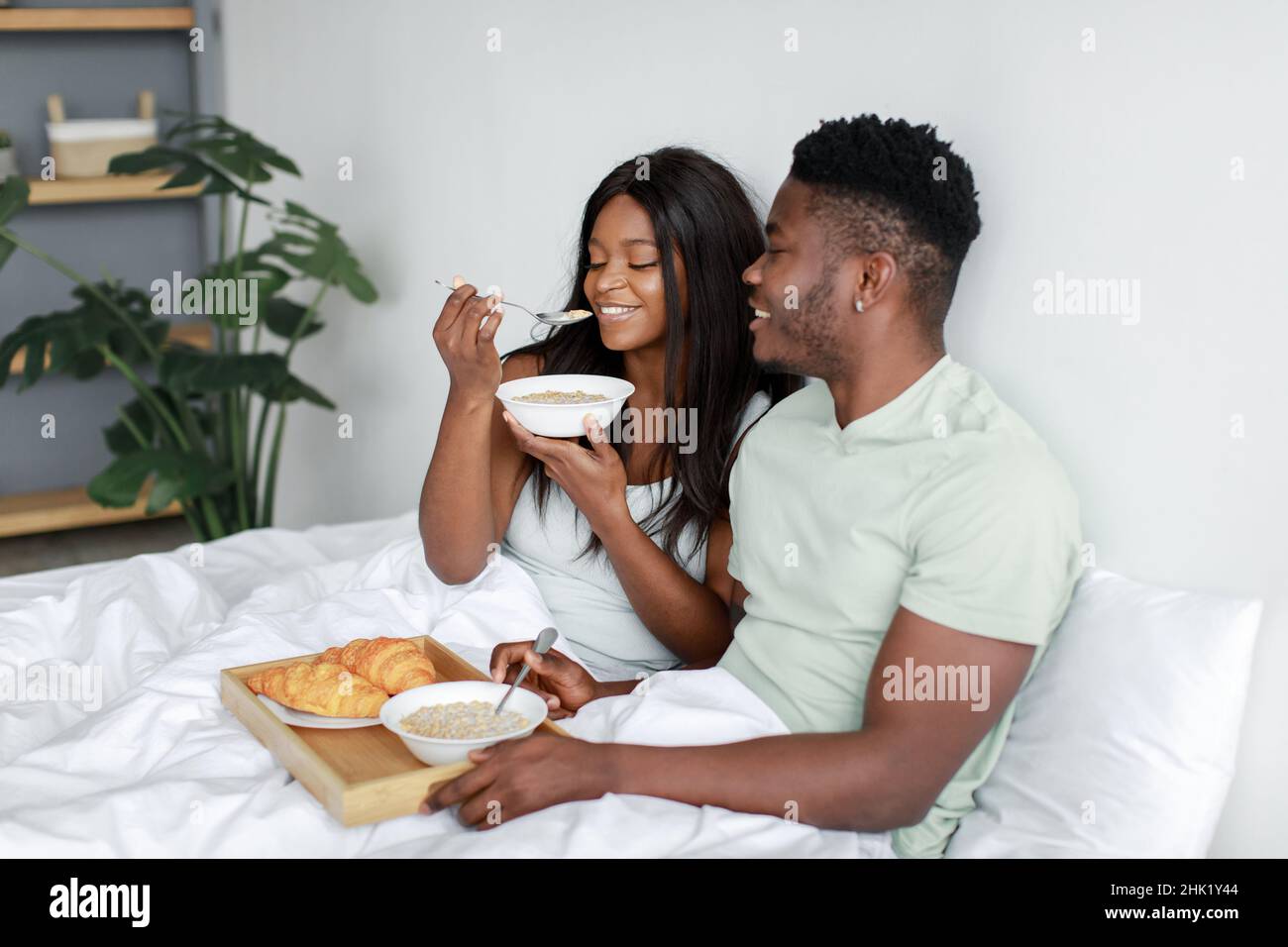 Glückliche, tausendjährige schwarze Frau und Ehemann essen Haferbrei auf weißem Bett im Schlafzimmer, genießen Sie das Frühstück und guten Morgen Stockfoto