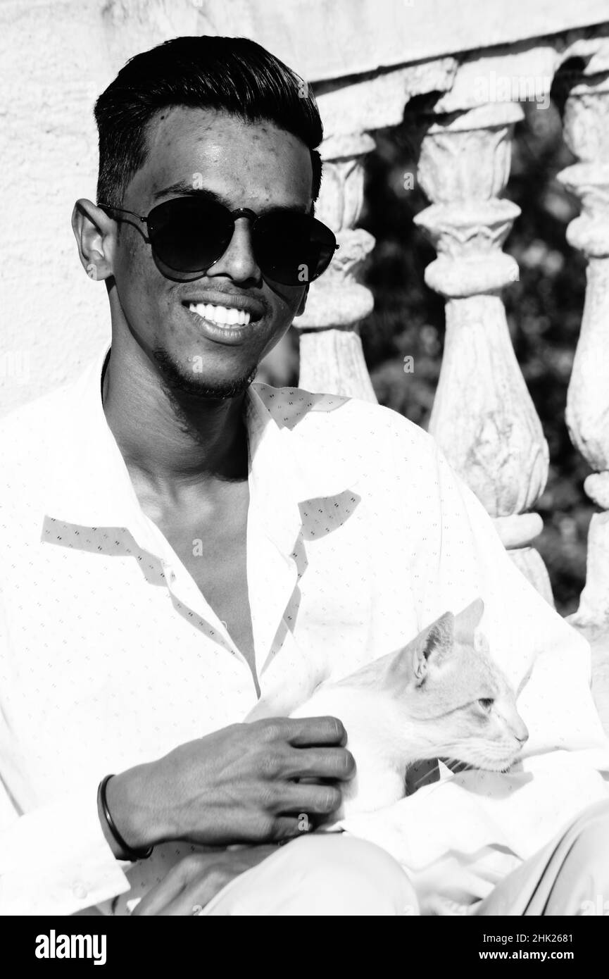 Graustufen eines südasiatischen Mannes in einer Brille, der mit einer Katze im Freien sitzt Stockfoto