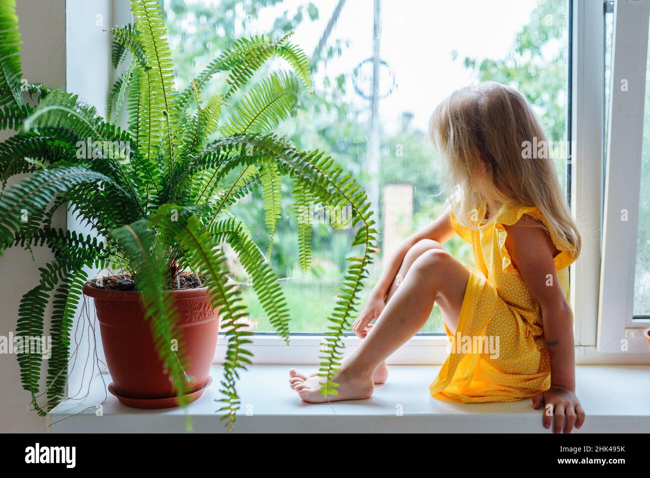 Porträt des blonden Mädchens barfuß sitzend auf Fensterbank wehmütig in der Nähe von Pflanzenfarn in Tontopf suchen. Seitenansicht. Stockfoto