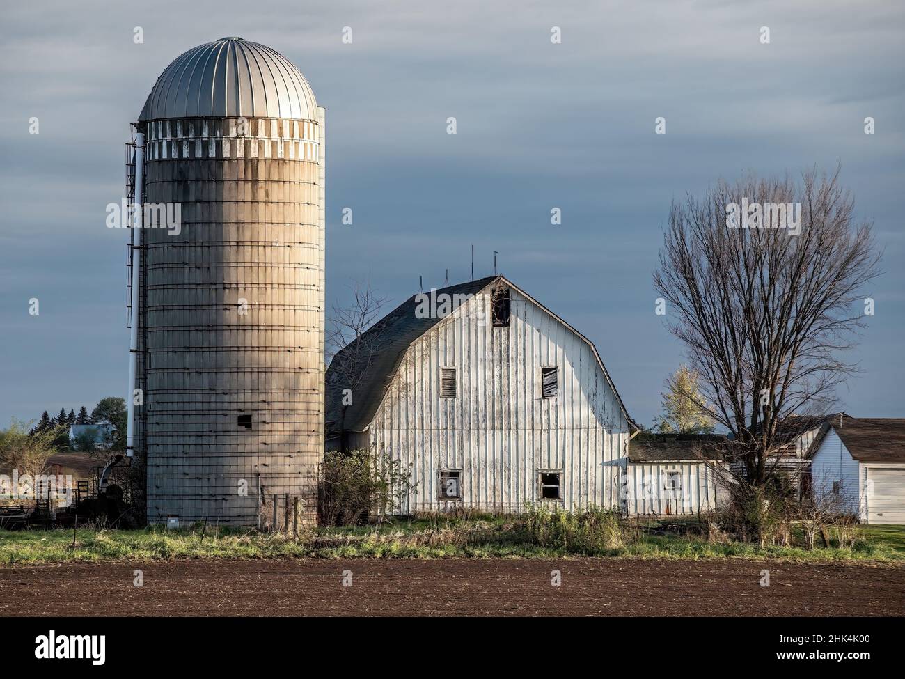 Farm mit einer weißen Scheune und Silo über ein Feld im Frühling bei Sonnenuntergang. Stockfoto