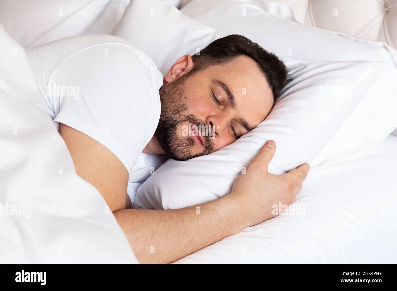 Hübscher junger Mann, der auf weißer Bettwäsche schläft. Blick auf einen gutaussehenden lächelnden Mann, der zu Hause in seinem Bett schläft. Stockfoto