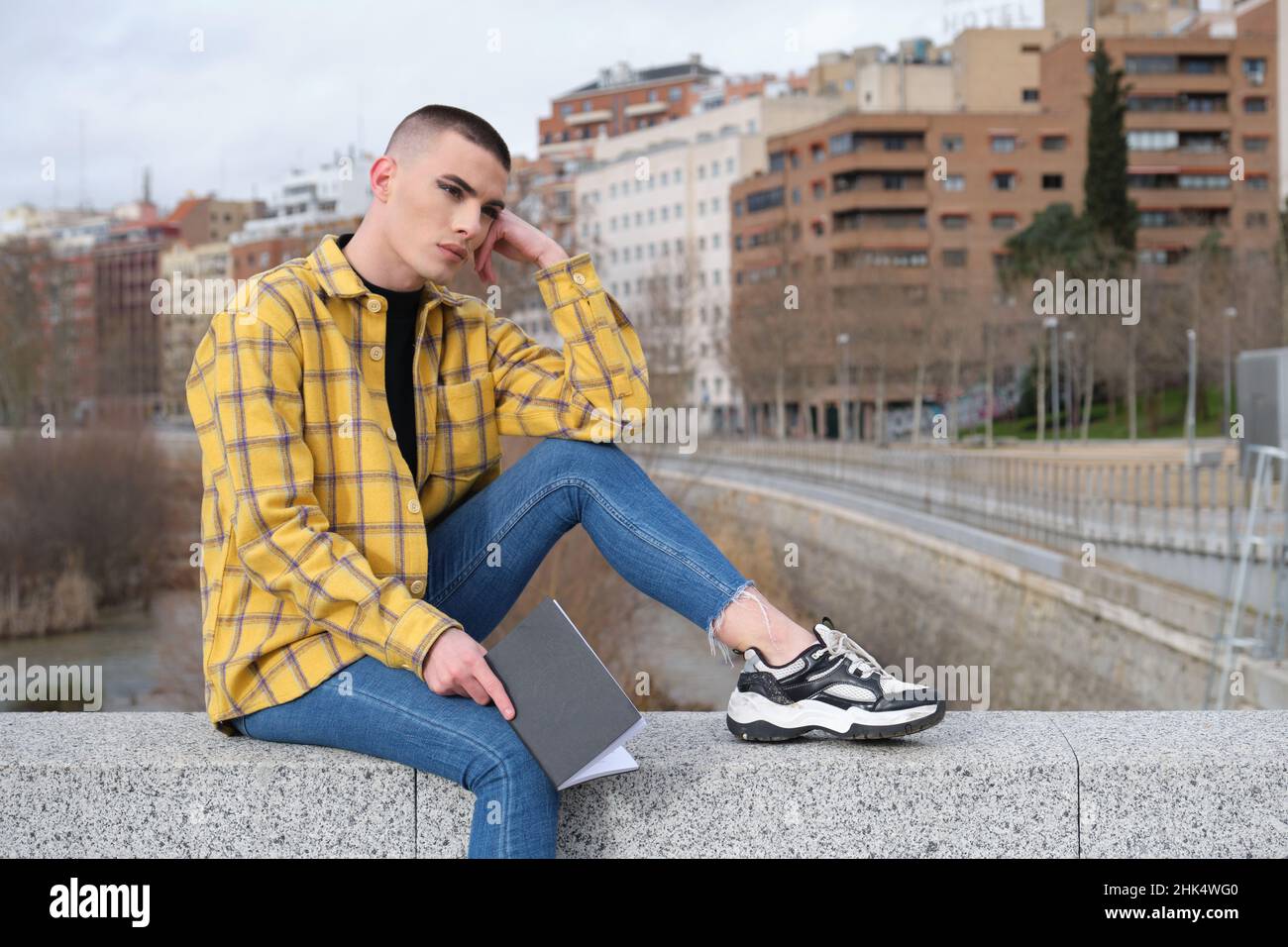 Nicht binäre Person, die auf einer Wand sitzt, mit einem Buch mit einem leeren Blick auf seinem Gesicht. Stockfoto