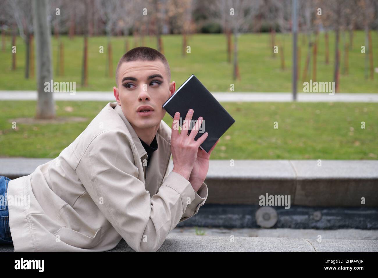 Junger Mann, der Schminke trägt und an einer Wand liegt und mit einem Buch zurückblickt. Stockfoto