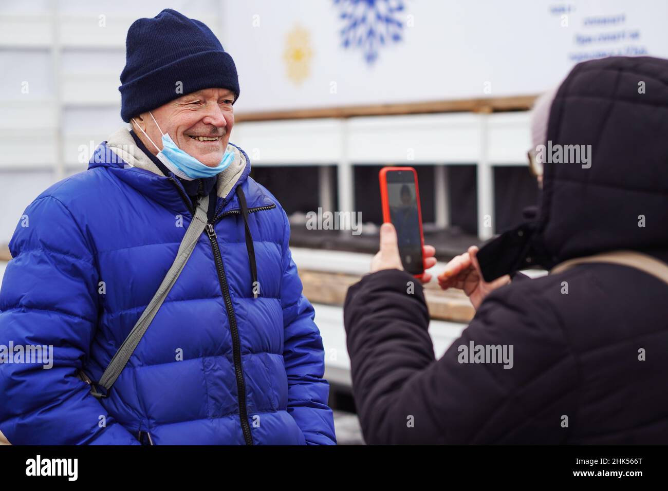 Ältere kaukasische Frau, die ihren grauhaarigen lächelnden Mann mit einer schützenden Gesichtsmaske fotografierte, älterer Mann, der am Smartphone hing. Senioren mit neuem Technol Stockfoto