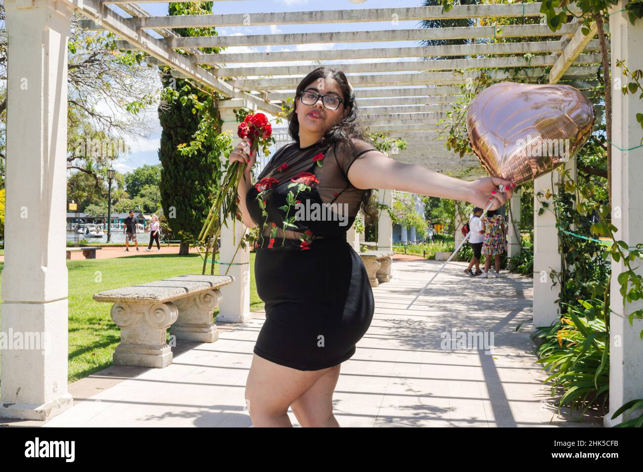 Das junge kaukasische lateinamerikanische Mädchen genießt den Valentinstag in einem Park, sie tanzt und hält einen herzförmigen Ballon und eine Blume in der Hand Stockfoto