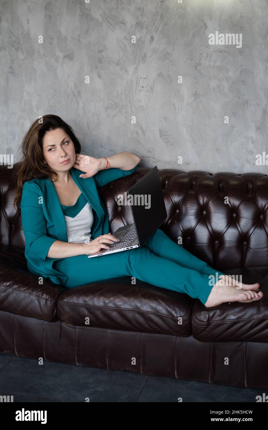 Fernarbeit, Fernstudium. Selbstbewusste Brünette Frau im grünen Anzug barfuß auf Ledercouch mit Laptop. Home Office. Arbeit von zu Hause aus. Stockfoto