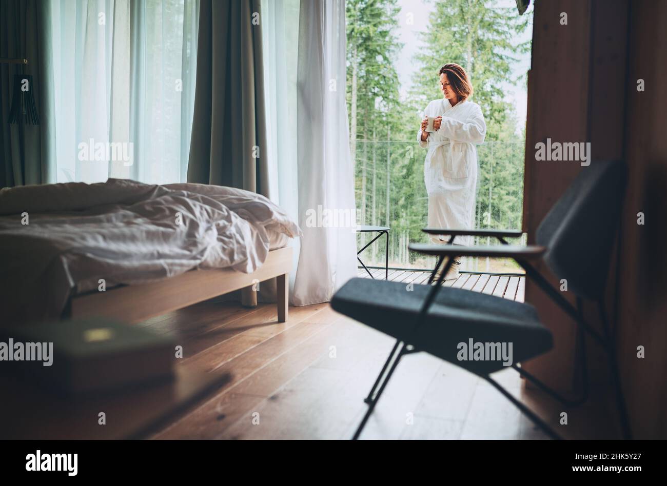 Frau gekleidet weißen Bademantel stehen auf Waldhaus Balkon und genießen frische Luft und Morgen Teetasse. Im Inneren eines skandinavischen Interior Design-Zimmers vi Stockfoto