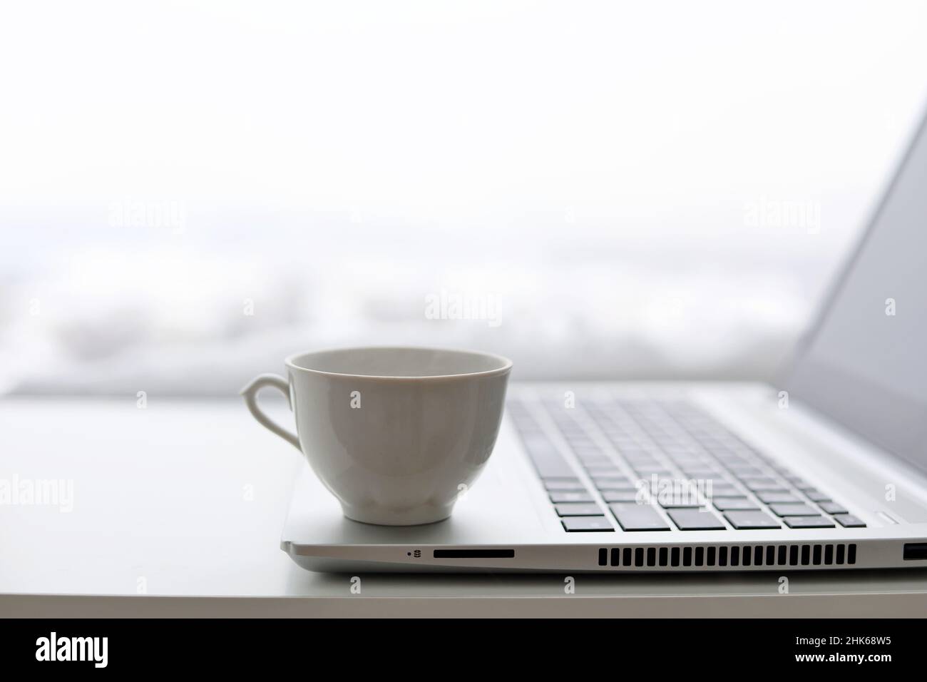 Kaffeetasse und Laptop auf dem Tisch gegen das Fenster, Blick auf die Winterstadt. Gemütlicher Arbeitsplatz im Home Office Stockfoto