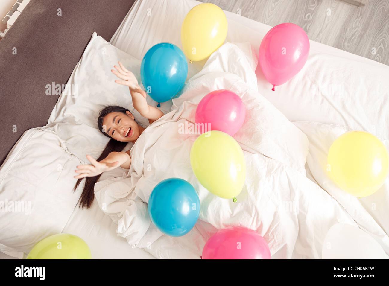 Fröhliche asiatische Frau im Bett mit bunten Ballons zu Hause liegen Stockfoto