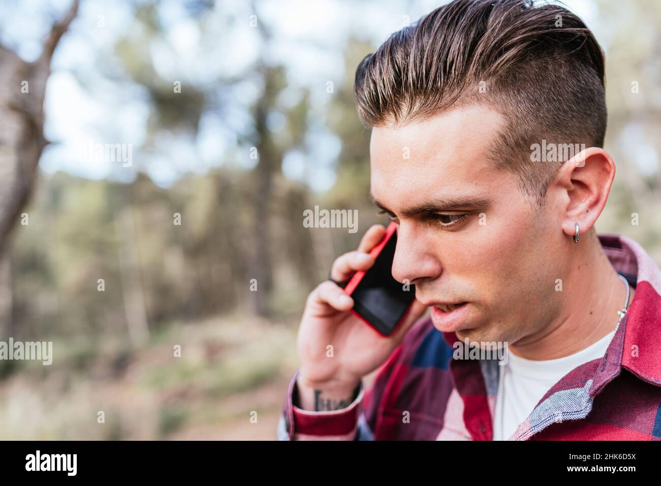 Erwachsener Mann im karierten Hemd, der auf dem Smartphone spricht, während er zwischen Bäumen im Hain steht Stockfoto