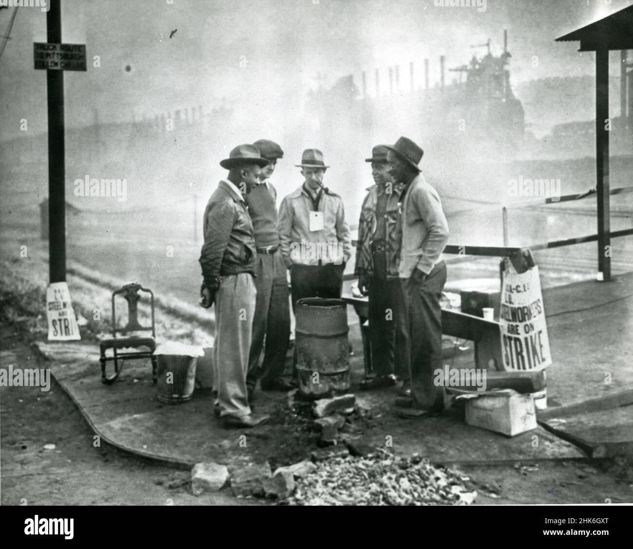 1949. Oktober, Duquesne, Pennsylvania - streikende Stahlarbeiter versammeln sich um ein kleines Feuer in einer alten Öltrommel, während sie sich die Zeit nehmen, die Bessemer-Fabrik der Carnegie-Illinois Co. Zu pickern.die Männer haben eine Bank, einen Schaukelstuhl und ein Radio an einer Straßenecke für ihre Mahnwache aufgestellt. Die Bessemer-Öfen im Hintergrund sind von Nebel umhüllt. Stockfoto