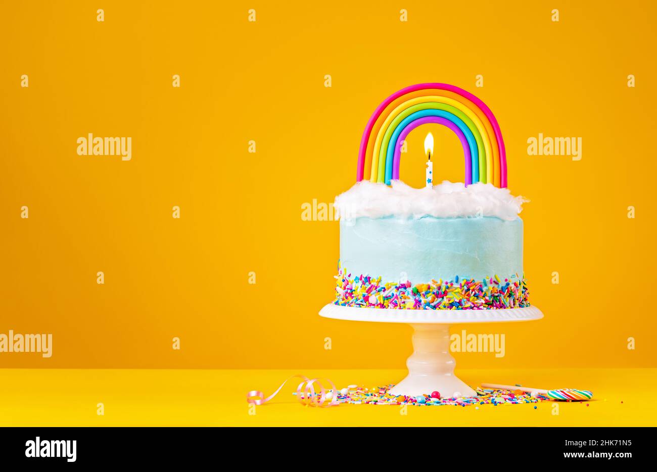 Blauer Geburtstagstorte mit Baiser-Dekorationen, Streuseln und einem farbenfrohen, hausgemachten Regenbogentopfer auf einem fröhlichen gelben Hintergrund. Stockfoto