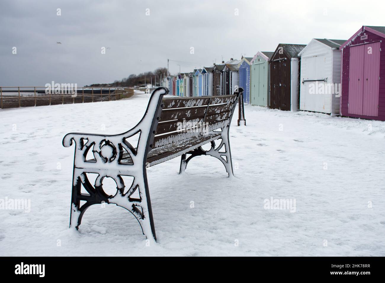 Schnee bedeckt das Ende einer öffentlichen Bank und bunte Strandhütten entlang der Promenade in Harwich & Dovercourt. North Essex, Großbritannien Stockfoto