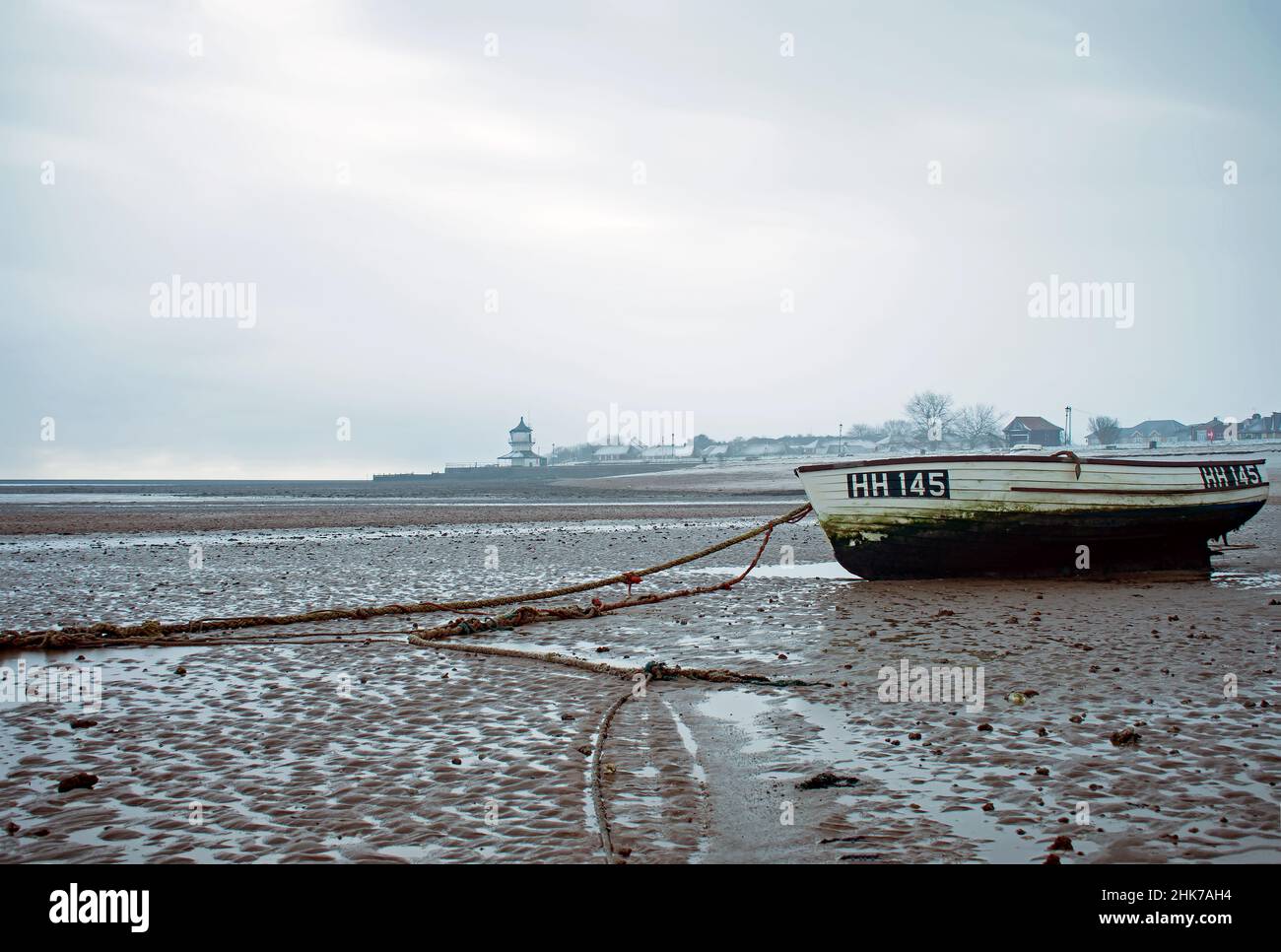 Bei Ebbe ruht auf dem Sand ein einflüchtiges Ruderboot. Harwich Low Lighthouse und Maritime Museum können im Hintergrund während des Winters Essex, Großbritannien, gesehen werden Stockfoto