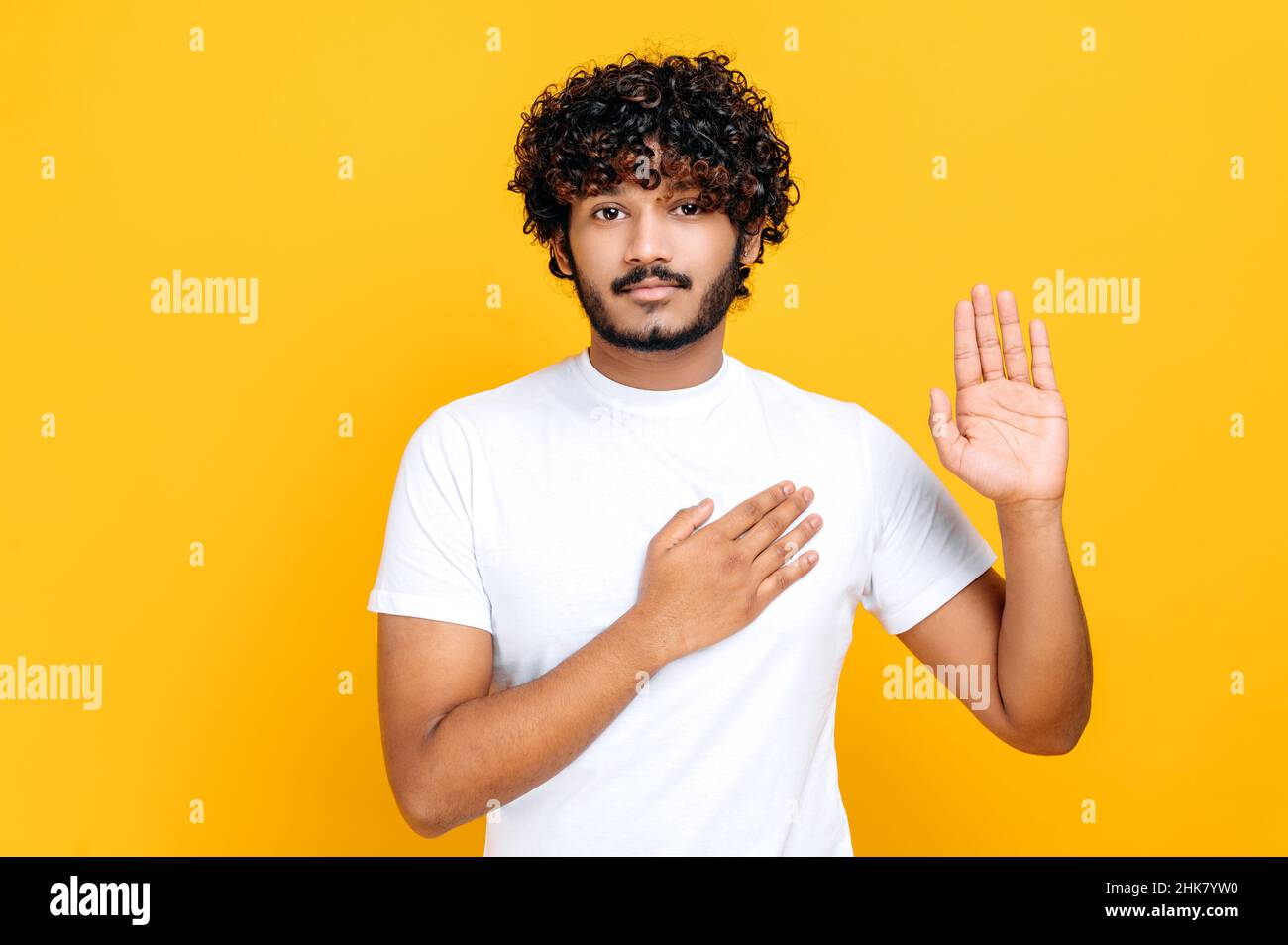 Ehrlicher, stolzer, lockiger junger, erwachsener Inder oder Araber in weißem, lässigem T-Shirt, die Hand auf der Brust und die Handfläche hebend, verheißend, eidend, auf isoliertem orangefarbenem Hintergrund stehend Stockfoto